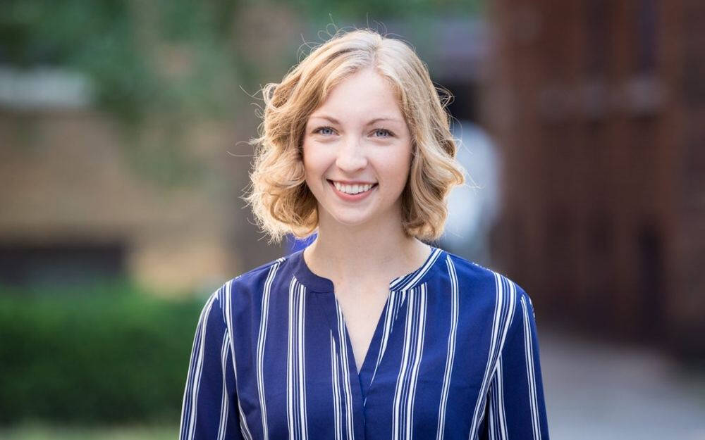 A smiling woman with short blonde curly hair wearing a blue and white striped blouse, standing outdoors with a blurred background.